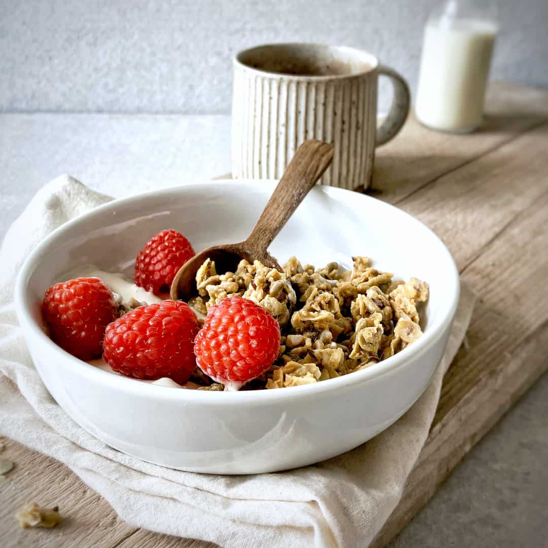 Raw Gorilla Peanut Butter Granola 2 Raw Gorilla Peanut Butter Granola in a white bowl with greek yoghurt and raspberries, with a wooden spoon, shot from the side.
