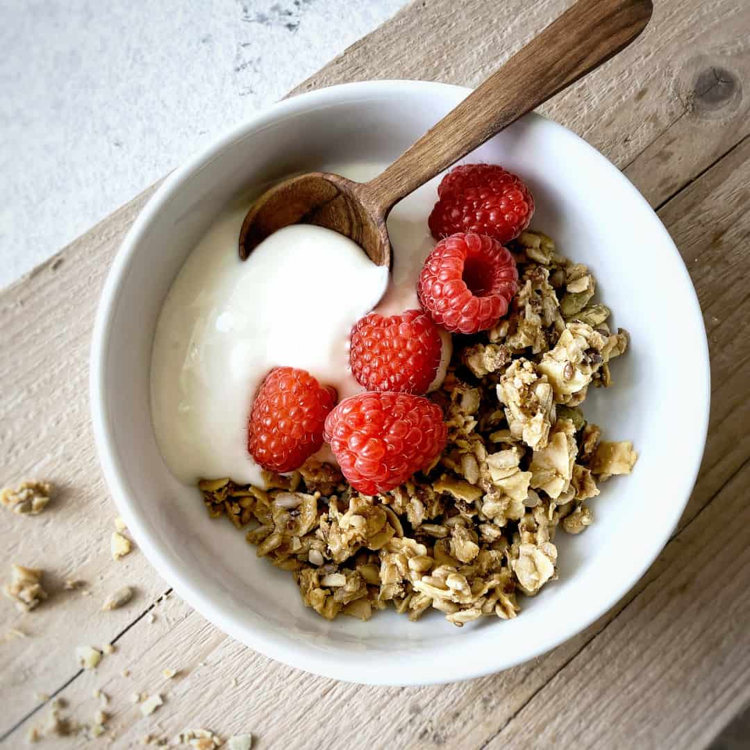 Raw Gorilla Peanut Butter Granola 3 Raw Gorilla Peanut Butter Granola in a white bowl with greek yoghurt and raspberries, with a wooden spoon, shot from above.