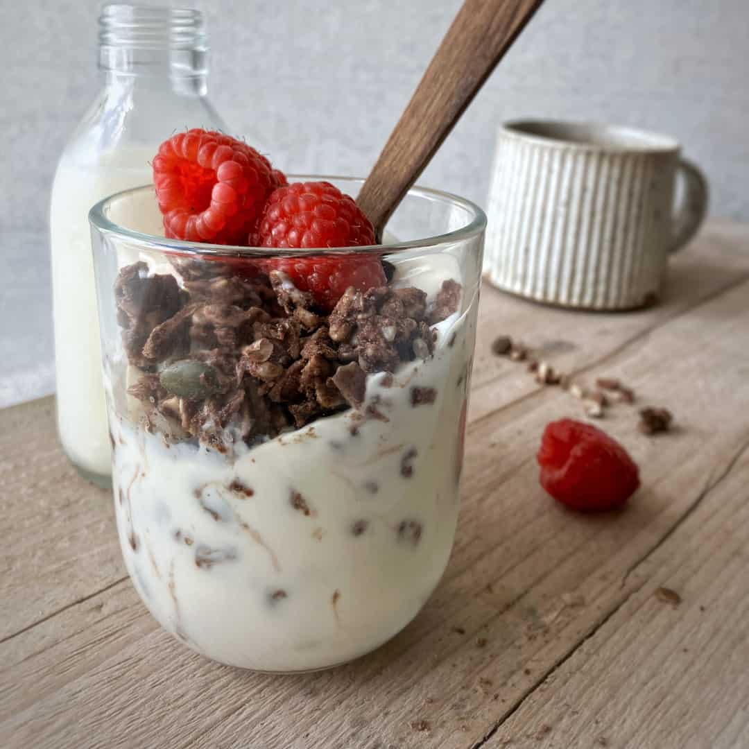 Raw Gorilla Peanut Butter and Chocolate Granola 2 Raw Gorilla Peanut Butter and Chocolate Granola in a glass with almond milk and raspberries, shot from the side.