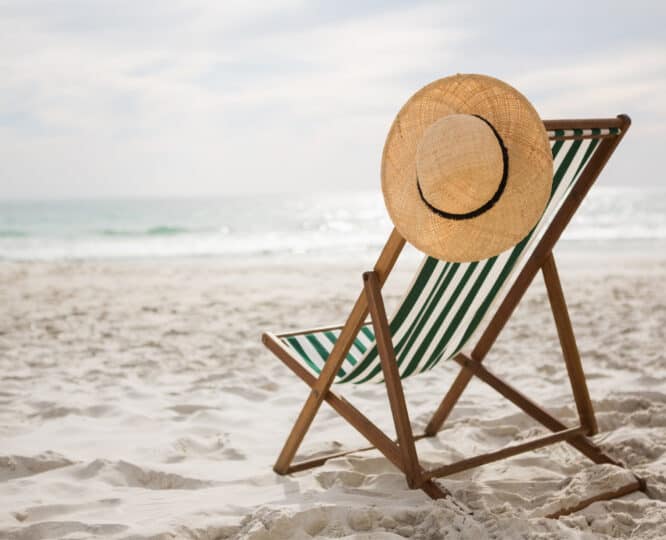 a beach char on a sandy beach with a sun hat resting on the top.